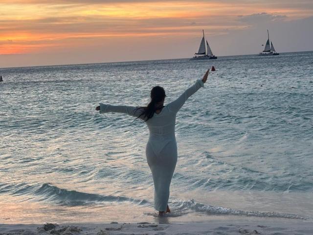 VictoriaWaynes with her ass out, arms spread, wearing a white dress, standing in shallow ocean water at sunset, with three sailboats in the background.