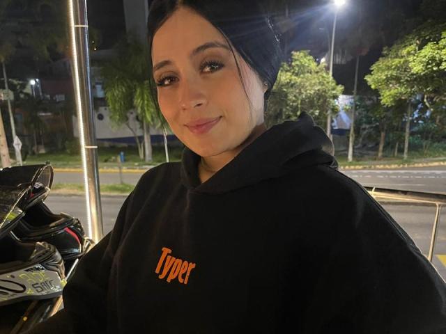 AnnyBentleii, avec des cheveux foncés, portant une capuche noire et le logo "Typer", avec un léger sourire, debout près d'une moto la nuit, éclairée par des lampadaires, avec des arbres verts en arrière-plan..