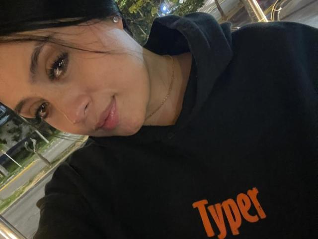 AnnyBentleii, avec ses cheveux foncés, ses yeux marron et ses lèvres roses, porte un sweat à capuche noir "Typer" et sourit légèrement. Son petit nez est orné d'un piercing discret..
