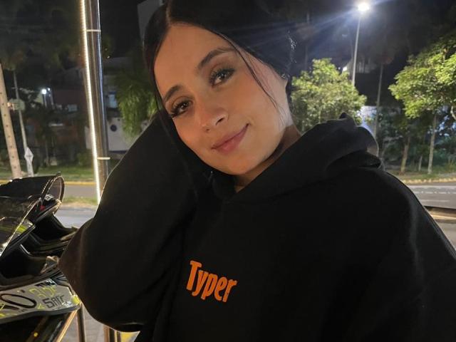 AnnyBentleii, avec ses cheveux noirs, porte un sweat à capuche noir "Typer", a un sourire charmant, une peau claire et tient ses cheveux relevés, se tenant près d'un vélo la nuit..