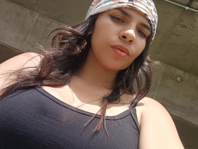 VioletaStone, met donker, golvend haar, draagt een cap met een patroon, een zwart tanktop, waardoor haar grote borsten zichtbaar zijn, en subtiele make-up. Ze kijkt in de camera met een lichte frons..