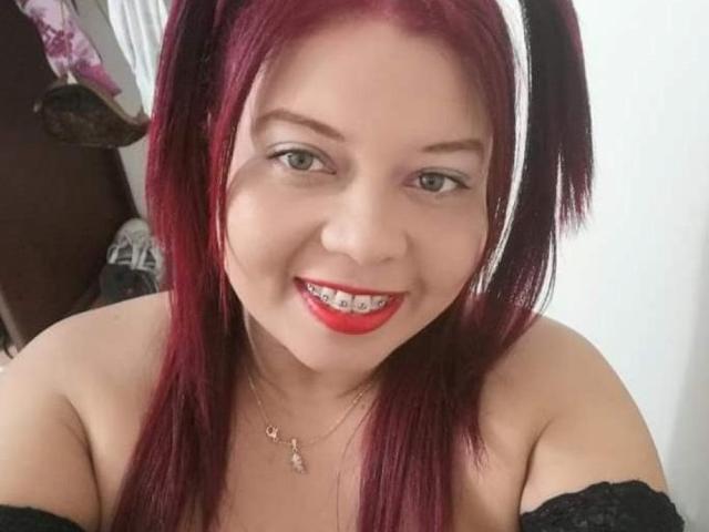 MilfAnalxx com cabelo ruivo, maria-chiquinhas, faixa de cabeça branca, batom vermelho, dentes de metal, sorrindo, blusa preta com um ombro descoberto, colar, pele clara, olhos verdes e um rosto fofo..