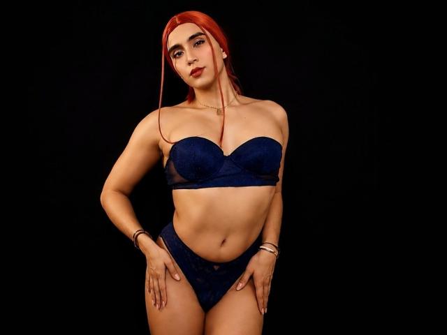 ClaryFrayx with red hair, wearing a blue lace bra and matching panties, standing against a black background, hands on her hips, looking confident, showing her tits and pussy.