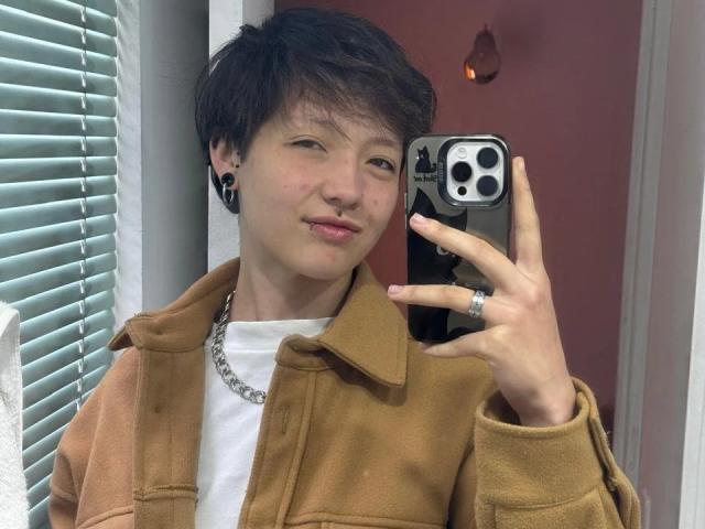 LissyLuxe with short brown hair, pink lips, wearing a brown jacket, white shirt, and black ear gauges, holding a phone, showing a ring, in a mirror selfie.