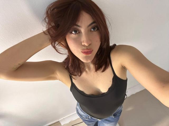 SpencerLe with brown hair, black lace-trimmed tank top, and blue jeans, looking up with pouty lips, hand in hair, standing in a white-walled room.