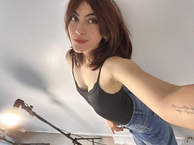 SpencerLe with red hair, black tank top, and blue jeans, showing a tattoo on her right arm, taking a selfie with a lamp in the background.