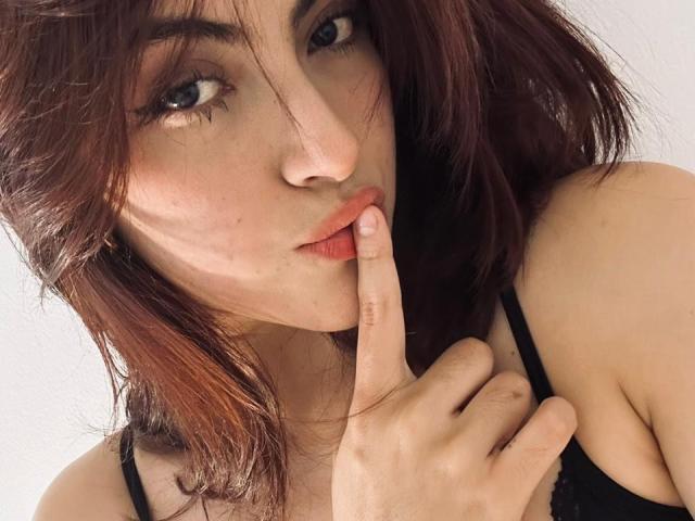 SpencerLe with dark red hair, pouty lips, and finger to mouth, wearing a black top, showing off her tits slightly with a seductive look, in a selfie.