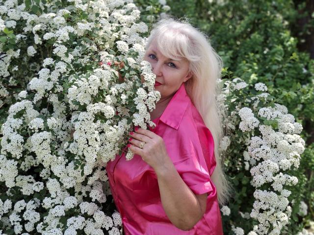KamaliaSim with long blonde hair, pink shirt, holding white flower bush, smiling, hands partially covering her tits, surrounded by greenery and white flowers, daytime, outdoors.
