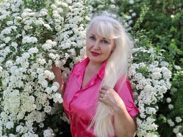 KamaliaSim with long, platinum blonde hair, pink shirt, and red lipstick, standing in a bush of white flowers, holding her hair with a ring on her finger.
