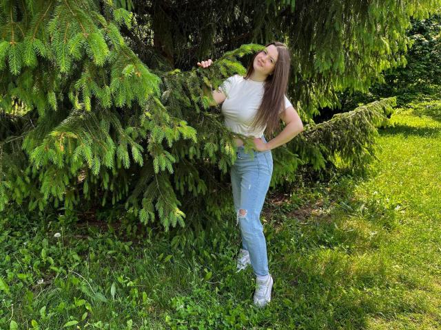 Mavka: Een meisje met lang bruin haar, een wit T-shirt, versleten blauwe jeans en witte sneakers, leunt tegen een groene dennenboom, glimlachend met haar handen op haar heupen..