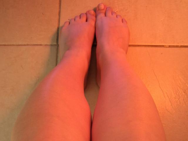 Marriam's bare feet with slightly pinkish skin, toes pointed upward, on a beige tiled floor, showing subtle shadows and slight red light reflection.