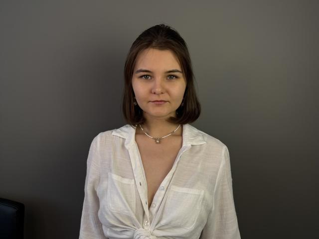 Nakisha Panchal, com cabelo castanho na altura dos ombros, vestindo uma camisa branca de botões, ligeiramente aberta, revelando seus pequenos seios macios e um colar de prata, está em pé contra um fundo cinza..