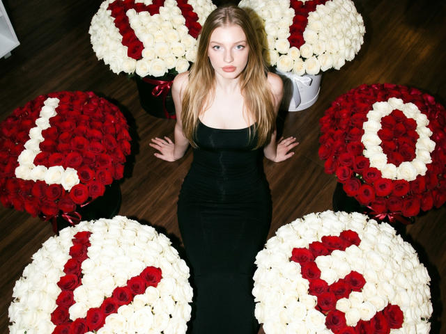 Ginnia with long blonde hair, wearing a black strapless dress, sitting among six round bouquets of red and white roses, on a dark wooden floor.