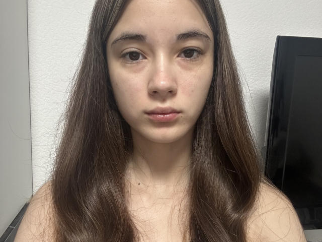 EleaseTichenor has long brown hair, pale skin, and brown eyes, with subtle makeup and a neutral expression. She's wearing a black top, and there's a TV in the background.