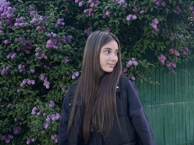 EleaseTichenor has long brown hair, wears a black jacket, and stands in front of purple lilac bushes with green leaves, looking to the side with a slight smile.