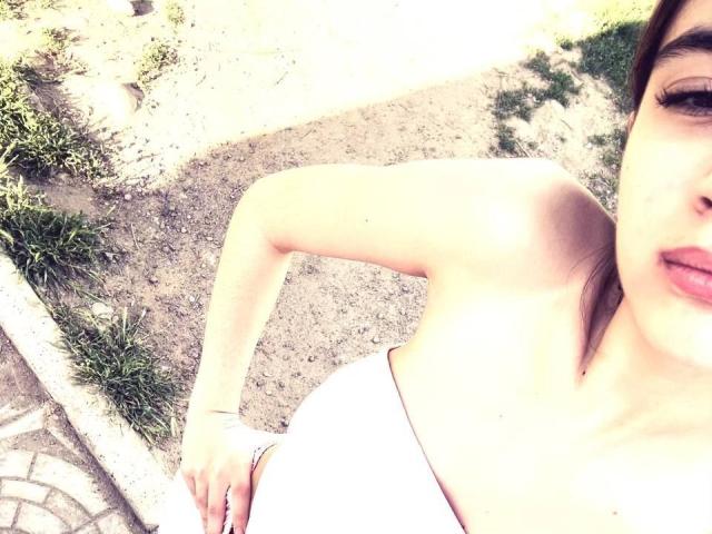 EleaseTichenor's tits are visible in a white strapless top, with her right arm raised and brown hair partially covering her face, standing on a gravel path.