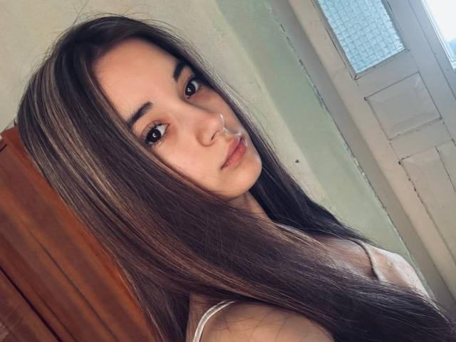 EleaseTichenor has long brown hair, brown eyes, and a small nose ring. She's looking at the camera with a slight pout, wearing a white top.