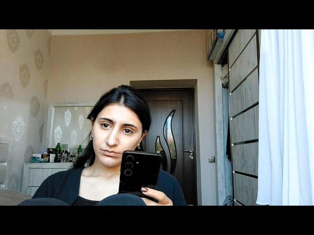 JaimeBorriello with dark hair, holding a phone, taking a selfie, looking serious, wearing a black top, showing a small, cute nose, and brown eyes, in a modest room.