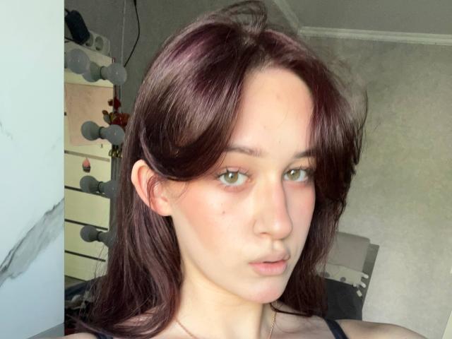 GarnetStelmack heeft een lichte huid, groene ogen en donkerbruin haar. Ze kijkt naar de camera met een neutrale uitdrukking, waarbij een kleine neus en lichtjes open lippen zichtbaar zijn..