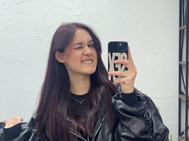 GarnetStelmack with long brown hair, winking, wearing a black leather jacket, holding a phone with a black and white case, smiling. Her tits are small, her ass is unseen.