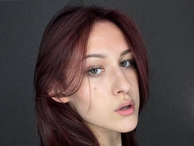 GarnetStelmack a les cheveux roux, les yeux verts et une petite tache de beauté sur sa joue droite. Ses lèvres sont légèrement entrouvertes, laissant entrevoir une expression subtile et séduisante..