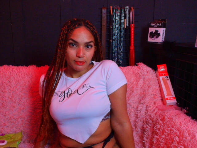 BrianaPratt with long braids, wearing a white crop top that shows her tits and a black thong, sitting on a pink fluffy couch with red lighting.