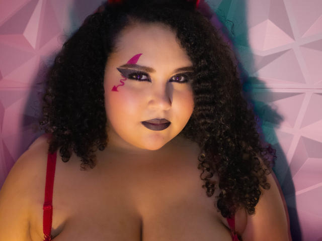 Curly-haired HurrenMatt with dark lipstick, red makeup lines, and red bra straps, showing her large boobs and confident gaze in a colorful, geometric-patterned photo.