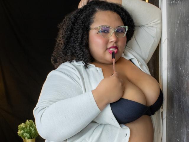 HurrenMatt with curly black hair, glasses, and red lipstick, holding a lipstick to her puffy tits in a black bra, wearing an open gray cardigan.