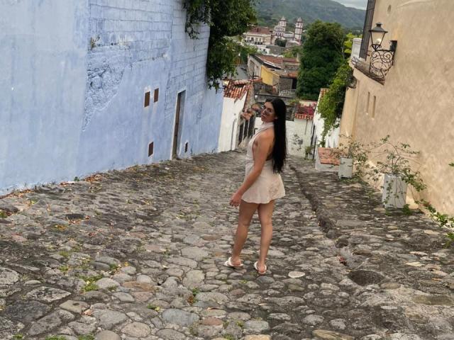 MiluSky stands on a stone pathway, wearing a short, lace, beige dress and white sandals, with long black hair, drinking from a bottle, showing her ass.