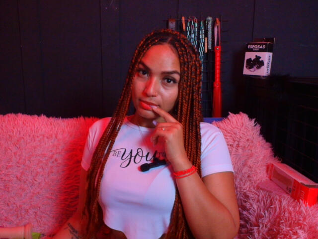 BrianaPratt with long braids, finger to lips, white tee, "you" text, red bracelet, pink fuzzy couch, red light, looking sexy, slightly pouting, dark background.
