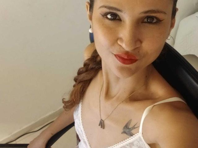 Amelia: Cabello rubio con labios rojos, blusa de encaje blanco, collar con placa de identificación, cabello castaño trenzado, tatuaje de pájaro en el pecho, mirando hacia arriba, sentada en una silla, delgada, busto mediano, piel clara..