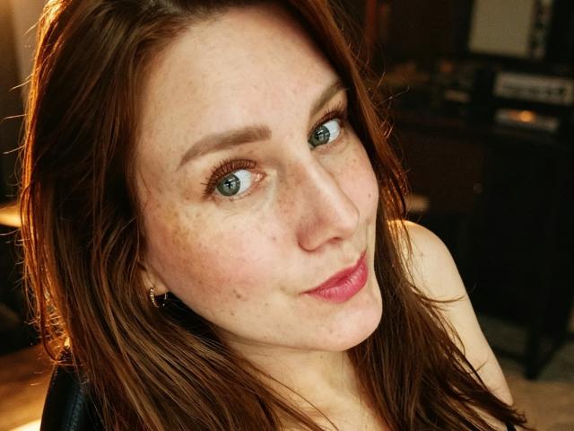 KatyaRogersX has blue eyes, freckles, and red lipstick with a slight smile. Her brown hair is loose, and she's wearing small hoop earrings. Her tits are not visible.