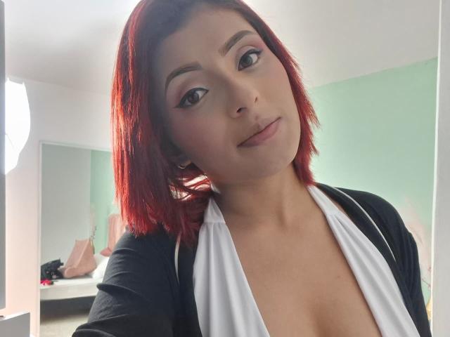 LizBonnet with red hair, white and black top, showing her tits in a selfie, green wall in background, subtle makeup, slight smile, looking at camera.