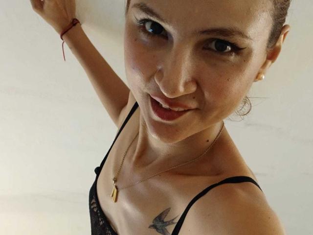 AmeliaBlond with a cute smile, black lace bra, small tits, tattoo of a bird on her chest, red bracelet, gold necklace, and earrings, raising her arm.