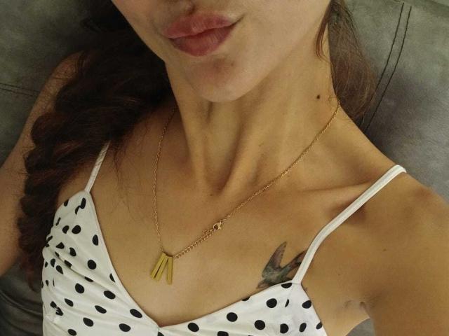 El pecho superior y el rostro de AmeliaBlond, mostrando un collar de oro con un dije en forma de "P", un vestido de lunares y un pequeño tatuaje de un pájaro en su pecho derecho..