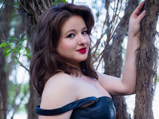 MarylinTaylor with wavy brown hair, red lipstick, and black off-shoulder leather top, leaning against a tree, showing her tits, looking at the camera with a slight smile.
