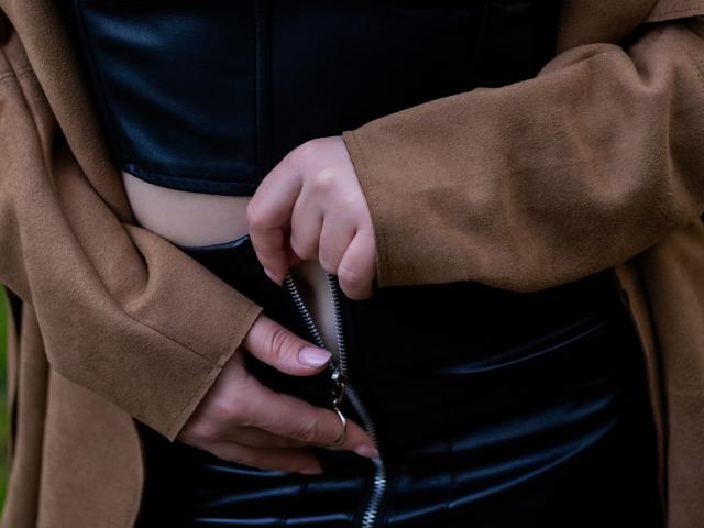 Marylin Taylor zieht mit ihren Händen schwarze Lederhosen an und trägt eine braune Wildlederjacke. Ihre Finger greifen den Reißverschluss, wobei ihre leicht blasse Haut sichtbar wird..
