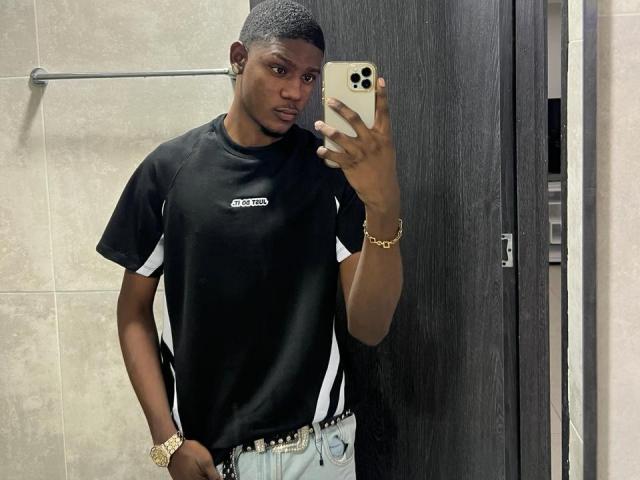 DrakeWit, donkerhuidig, met kort zwart haar, een zwart T-shirt met witte strepen, een gouden horloge, een gouden armband, vasthoudend een gouden iPhone, staand in een badkamer, kijkend in de spiegel..