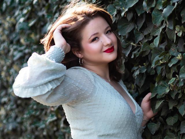 Marilyn Taylor con rossetto rosso, indossa un abito bianco trasparente con pois, si tocca i capelli castani, è in piedi di fronte a un muro verde con foglie, sorride dolcemente, mostrando leggermente il seno, appare carina..