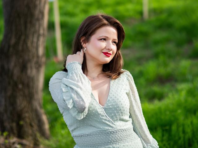 Marilyn Taylor, con senos de tamaño medio, vestida con un vestido verde, de corte profundo en V y con lunares, con lápiz labial rojo, cabello castaño, con la mano tocándose la oreja, de pie junto a un árbol en un área verde y cubierta de hierba..