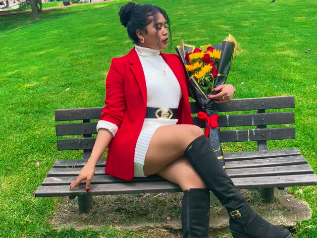 SophieSonyas sitting on a bench, wearing a red jacket, white turtleneck, and white mini-skirt, holding flowers, with black knee-high boots, and a red belt, looking to the side.