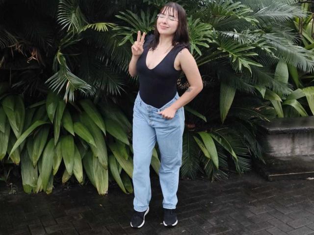 HairyValeri, con piccoli seni, indossa una canotta nera, jeans azzurri e scarpe da ginnastica nere, fa un gesto di pace, in piedi di fronte a una vegetazione verde e rigogliosa..