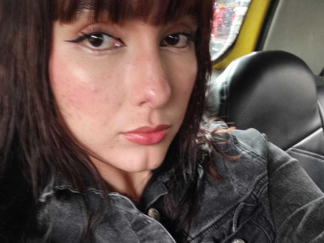 HairyValeri, con i capelli castani, la frangia e l'eyeliner nero, indossa una camicia di jeans grigia, ha il rossetto rosa e un'espressione leggermente corrucciata, ed è seduta in un'auto..