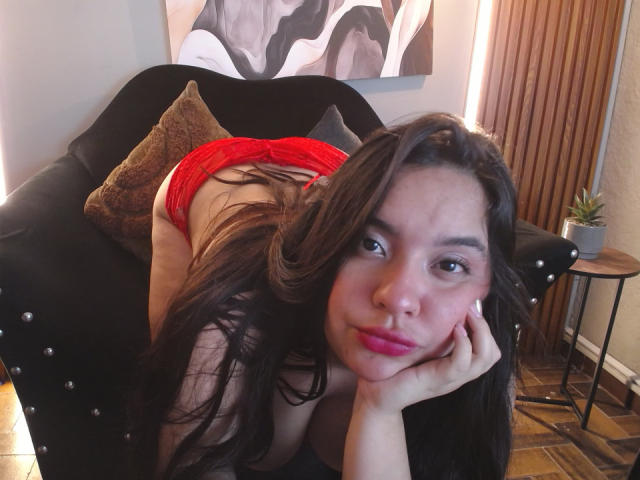 SammyTitss è seduta su un divano nero, indossa delle mutandine di pizzo rosse, è piegata in avanti con il sedere sollevato, ha lunghi capelli castani, rossetto rosa e un viso sexy e sicuro di sé..
