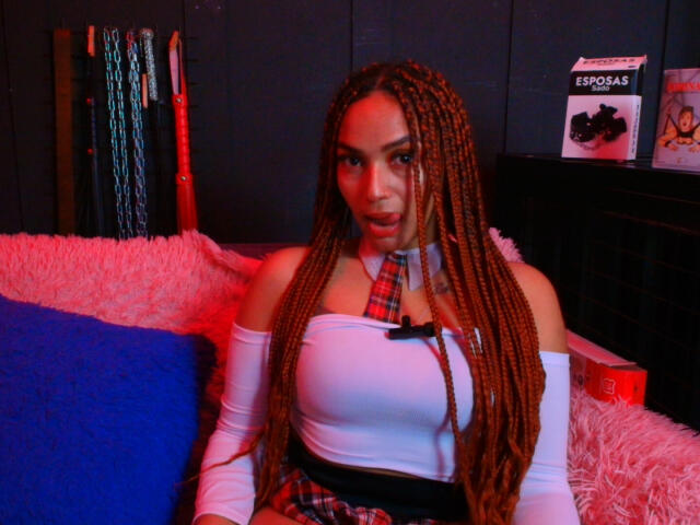 BrianaPratt with long braids, wearing a white off-shoulder top, plaid necktie, and matching skirt, sitting on a pink fluffy couch, with blue pillow, looking at the camera.