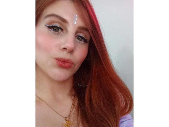 AngeliqueAnne has red hair, white face paint on her forehead, and is pouting with pink lips. She's wearing blue eyeliner and a gold necklace. Her tits are not visible.