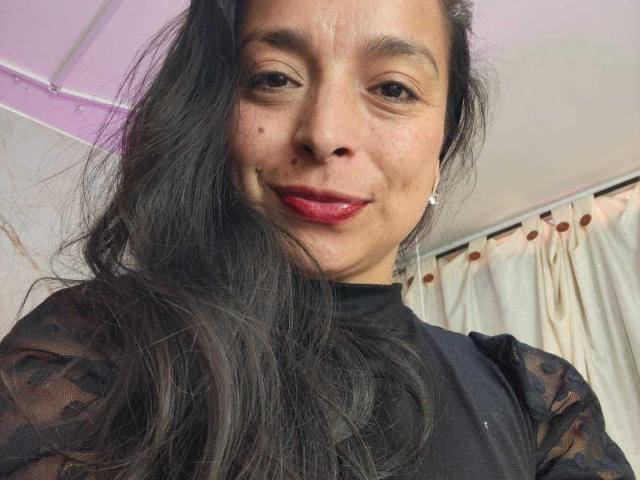 StangelarRivera has dark, wavy hair, red lipstick, and a black lace top, with a small mole on her right cheek, smiling at the camera.