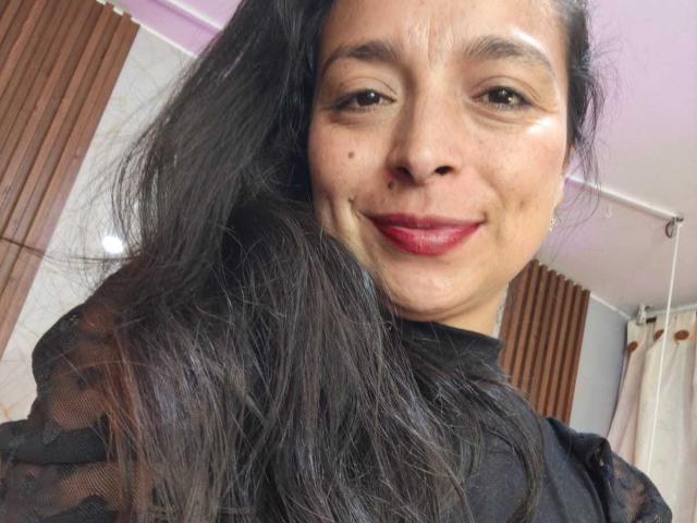StangelarRivera with long black hair, brown eyes, red lipstick, black sheer top, smiling at the camera, visible moles on cheek, medium skin tone, slightly messy hair.