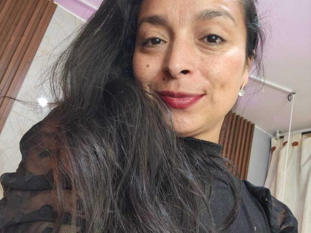 StangelarRivera has long black hair, brown eyes, and red lipstick. She's smiling with a small beauty mark on her left cheek, wearing a sheer black top.