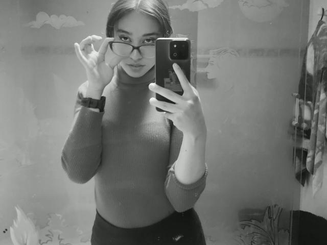 Autoportrait en noir et blanc de CamiVargas, portant des lunettes, un pull col roulé et une montre, tenant un téléphone, ajustant ses lunettes avec sa main gauche, montrant son visage et sa partie supérieure du corps, et portant un pantalon foncé..